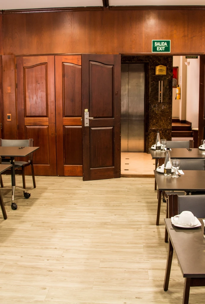 Sala de conferencias con mesas, sillas y salida hacia un ascensor.