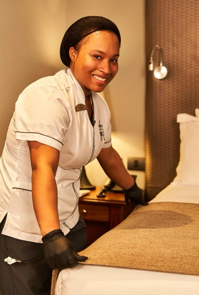 Mujer sonriendo mientras arregla la cama en una habitaci&oacute;n de hotel.