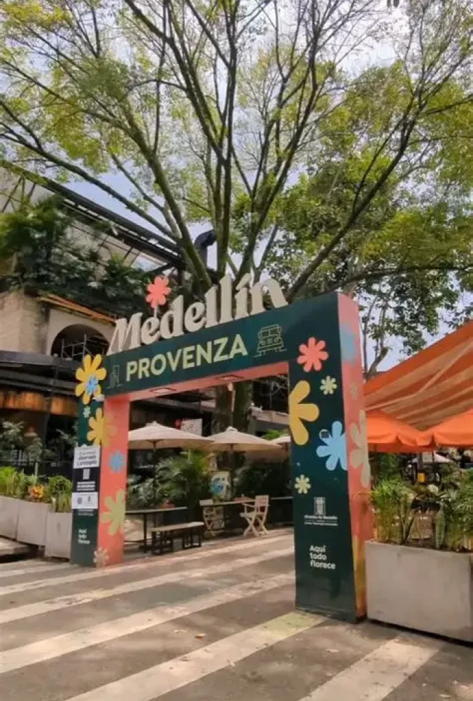 Entrada colorida al barrio Provenza, Medell&iacute;n, con &aacute;rboles y mesas al fondo.