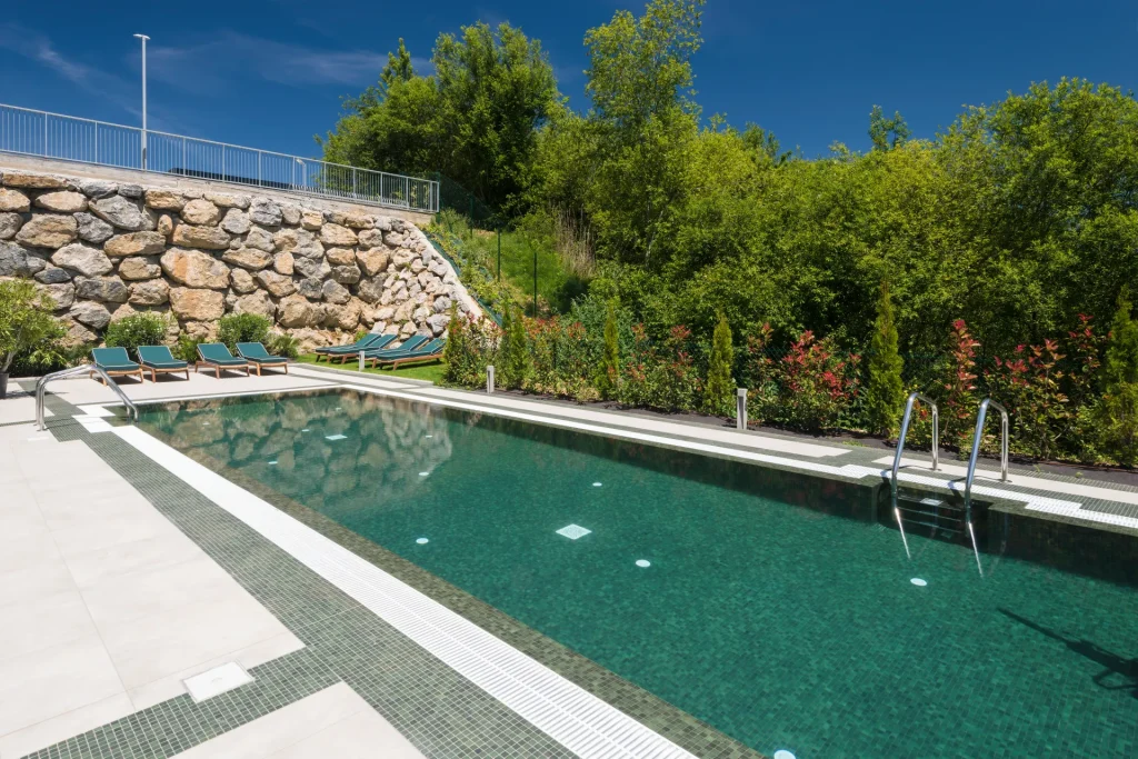 Piscina rodeada de vegetaci&oacute;n y tumbonas bajo un cielo azul claro.
