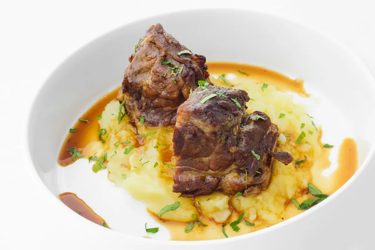 Carne estofada sobre pur&eacute; de papas y hierbas en un plato blanco.