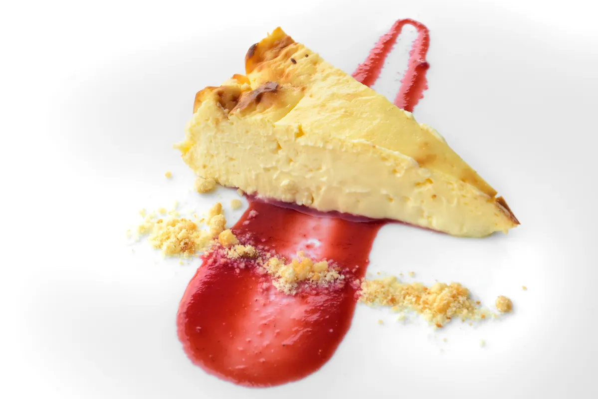 Trozo de tarta de queso con salsa roja y migas sobre un plato blanco.