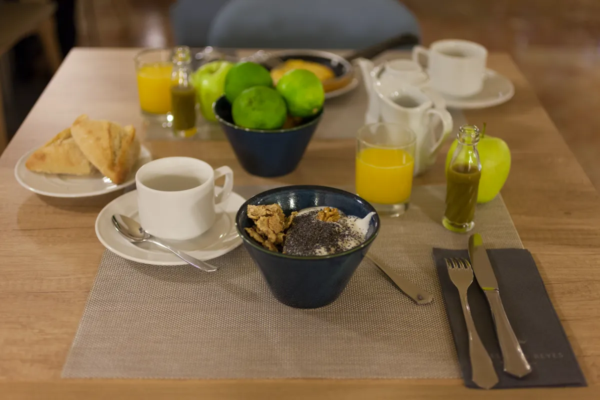 Desayuno con yogur, fruta, jugo, caf&eacute; y pan sobre una mesa.