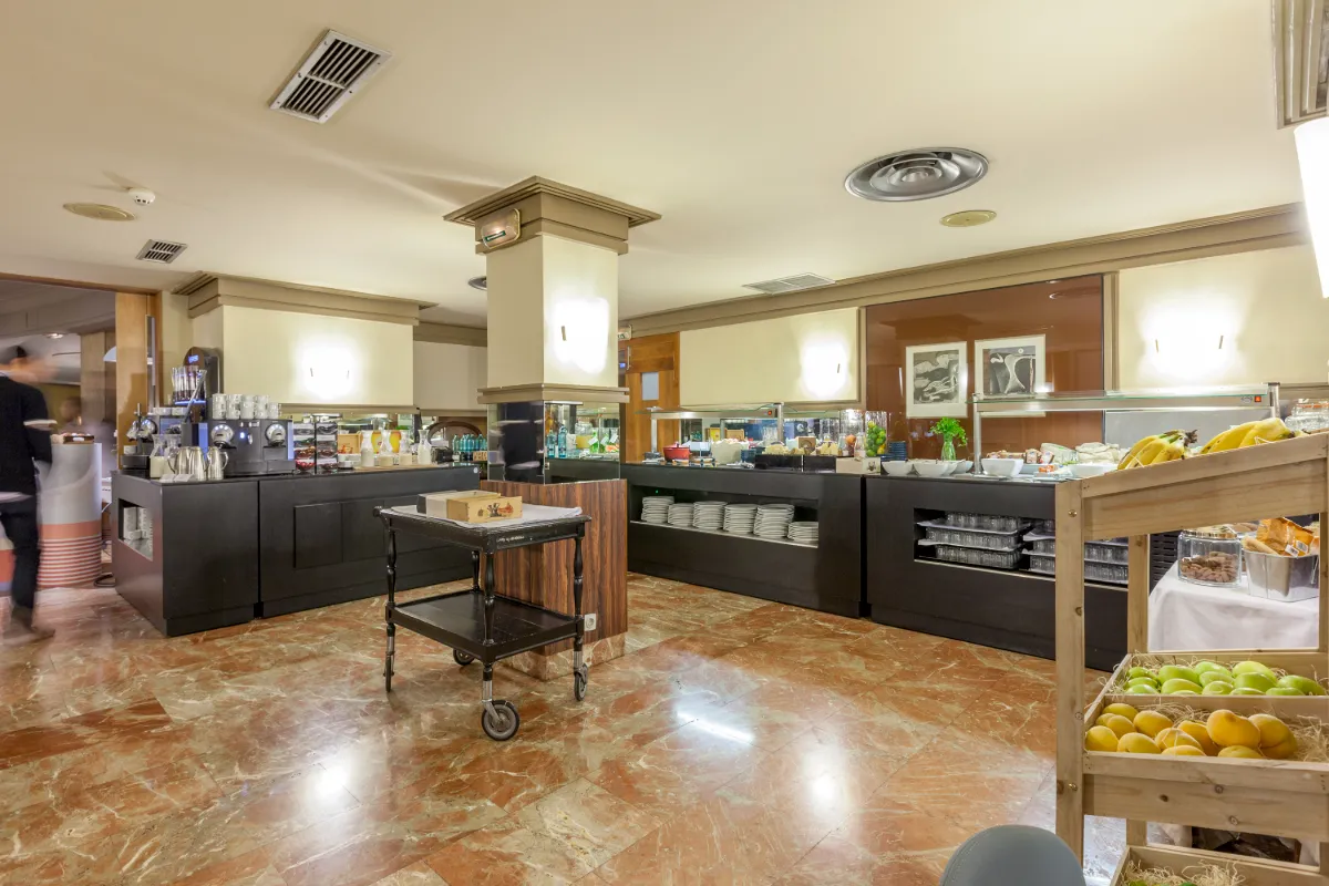 Buffet de desayuno con frutas, bebidas y utensilios en un hotel luminoso.