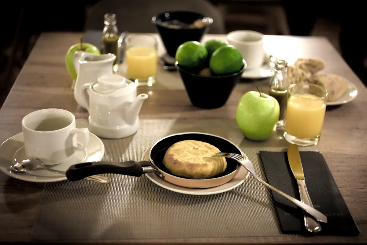 Desayuno con tortilla, t&eacute;, jugo y fruta en mesa de madera.