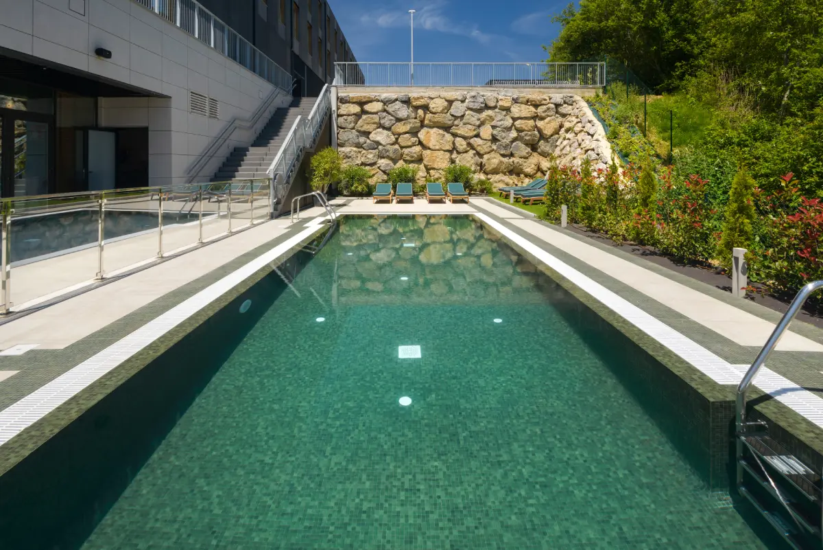Piscina al aire libre con tumbonas y jardines alrededor en un d&iacute;a soleado.