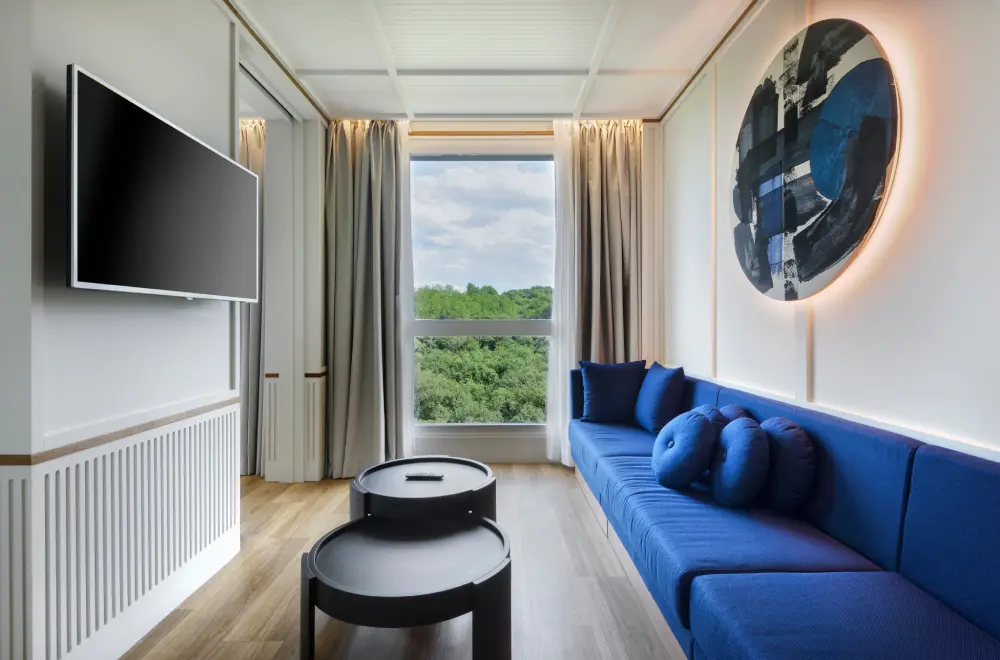 Sala moderna con sof&aacute; azul, mesa negra, televisor y ventana con vista al bosque.