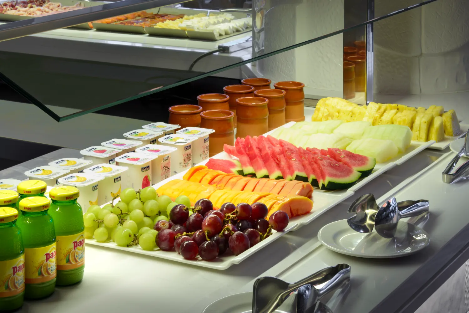 Buffet con frutas, yogures y jugos en una mesa con utensilios.