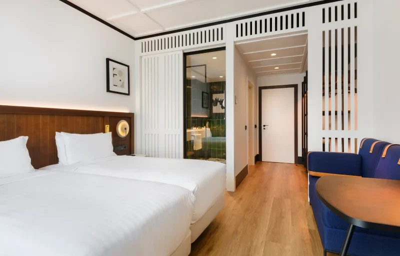 Habitaci&oacute;n de hotel moderna con dos camas, sof&aacute; azul y ba&ntilde;o visible.