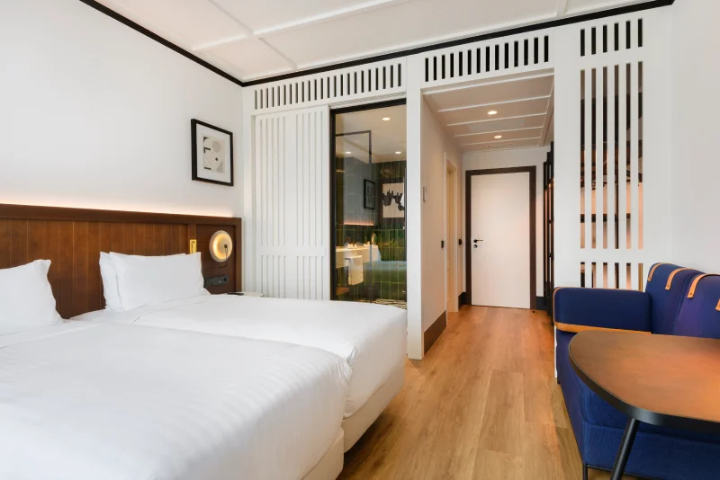 Habitación de hotel moderna con dos camas, sofá azul y baño visible.