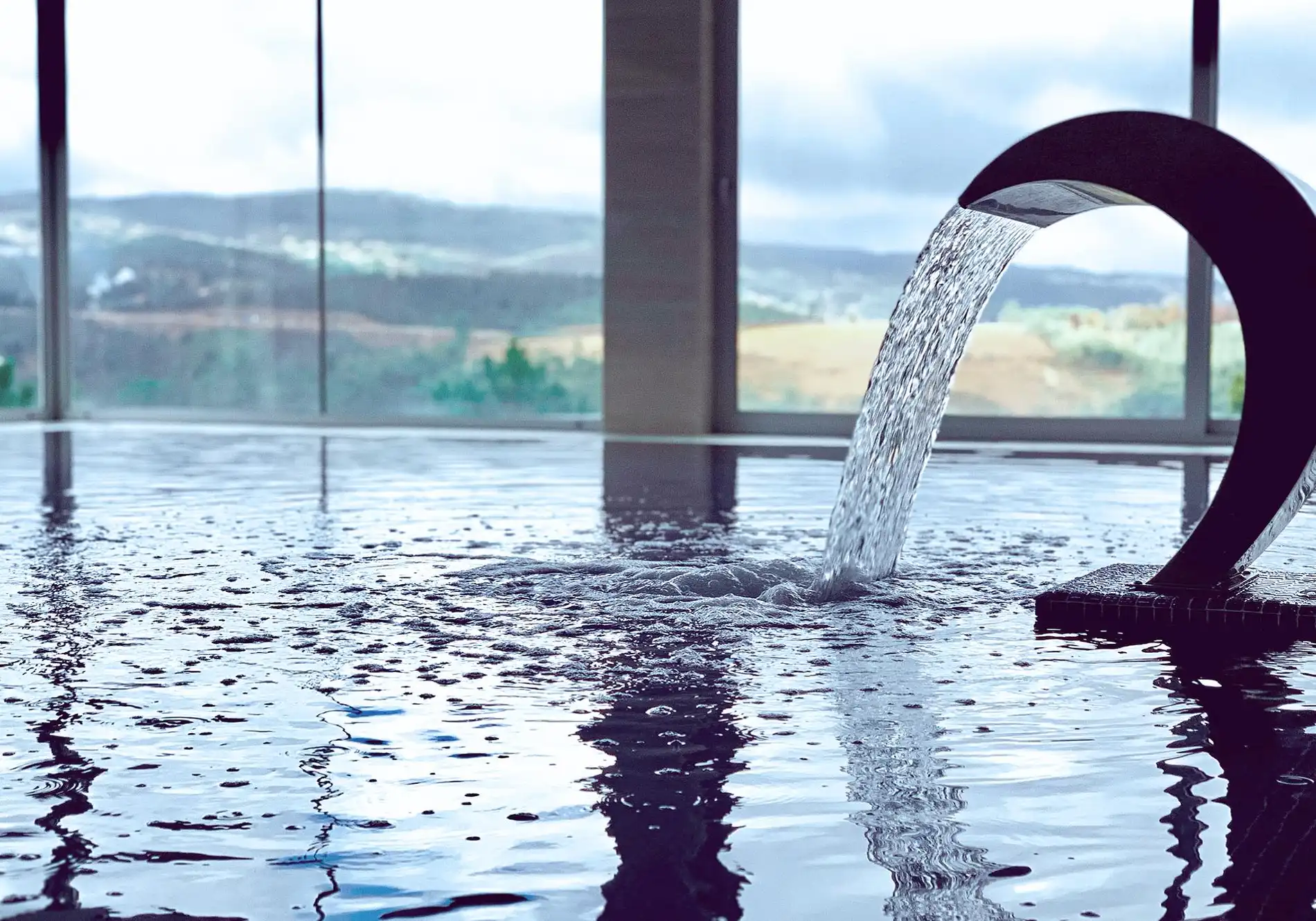 Fonte de &aacute;gua em piscina com janelas e vista para montanhas ao fundo.