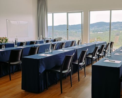 Sala de confer&ecirc;ncia vazia com mesas e cadeiras, vista para montanhas.