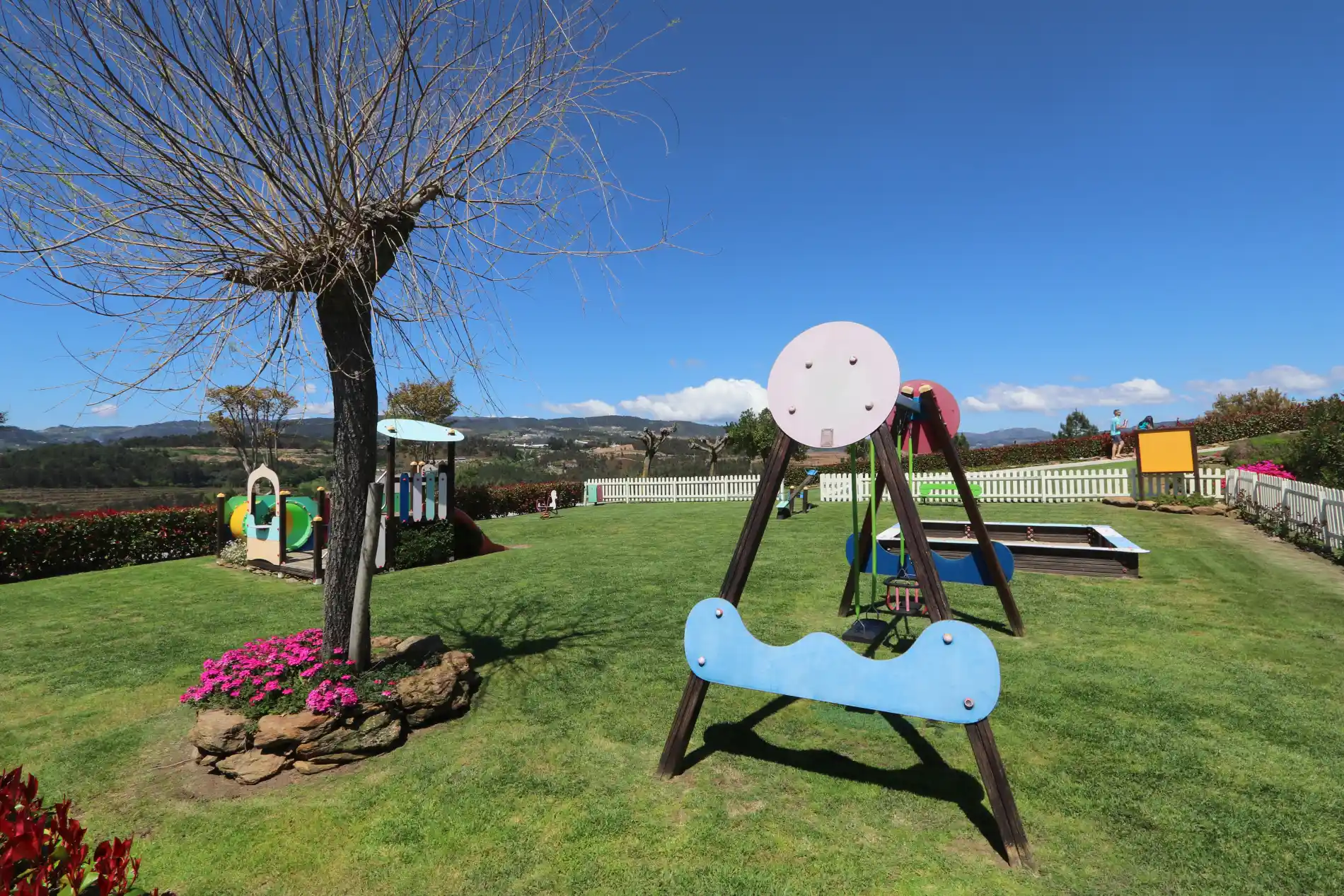 Parque infantil ao ar livre em dia ensolarado, com brinquedos e grama verde.