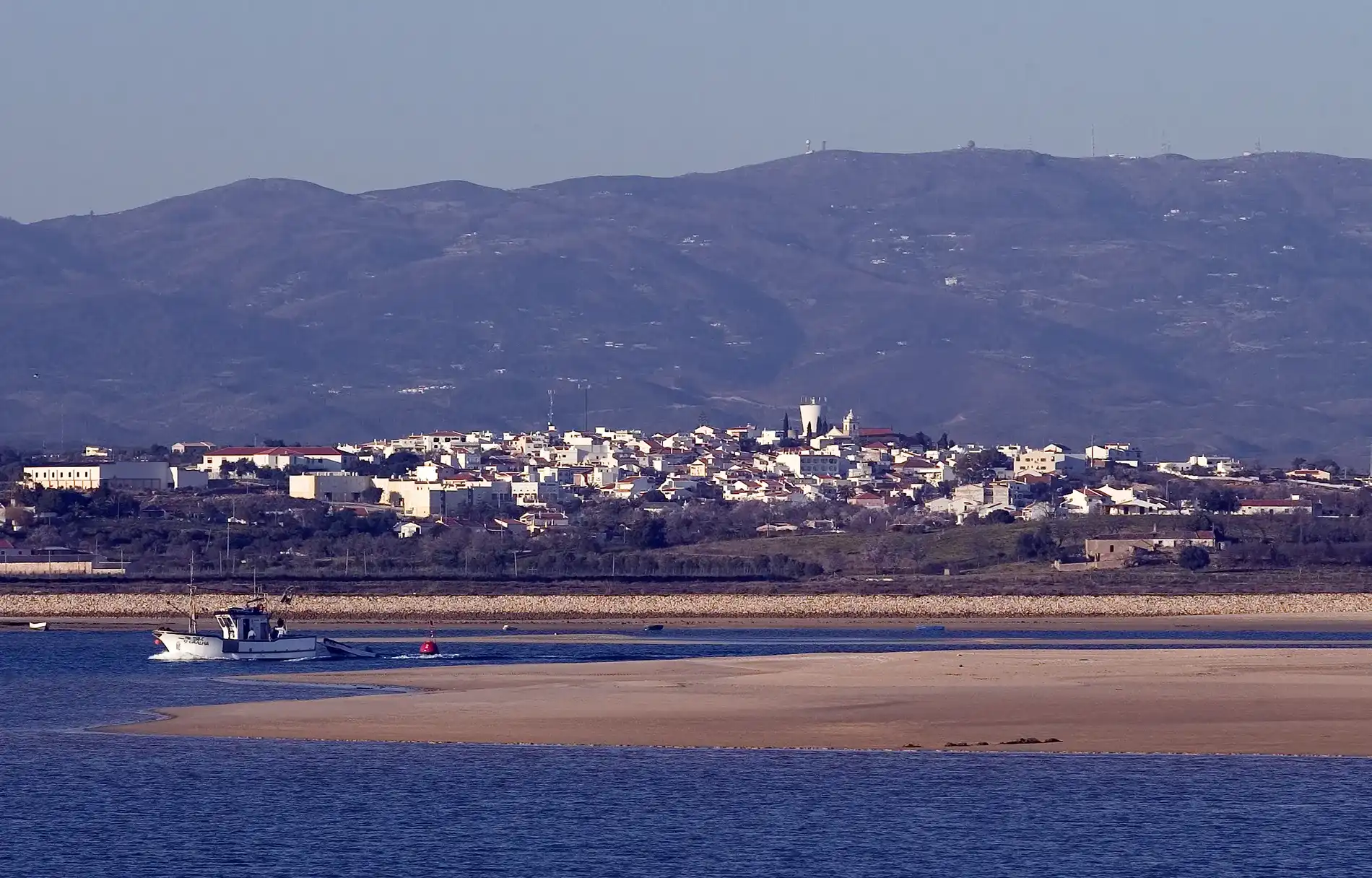 Vista de cidade litor&acirc;nea com montanhas ao fundo e barco no mar.