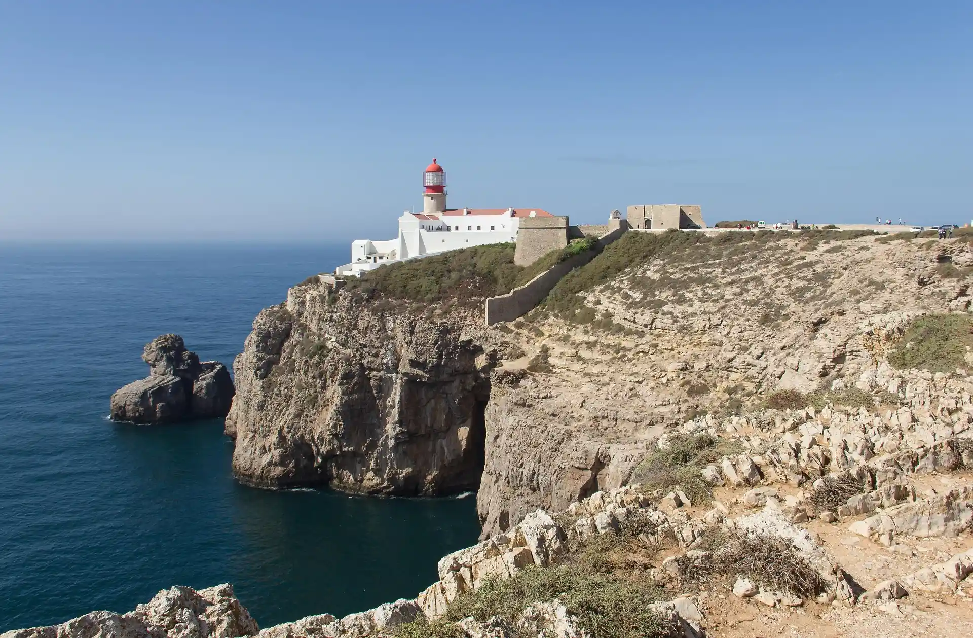 Farol em penhasco rochoso junto ao mar sob c&eacute;u azul.