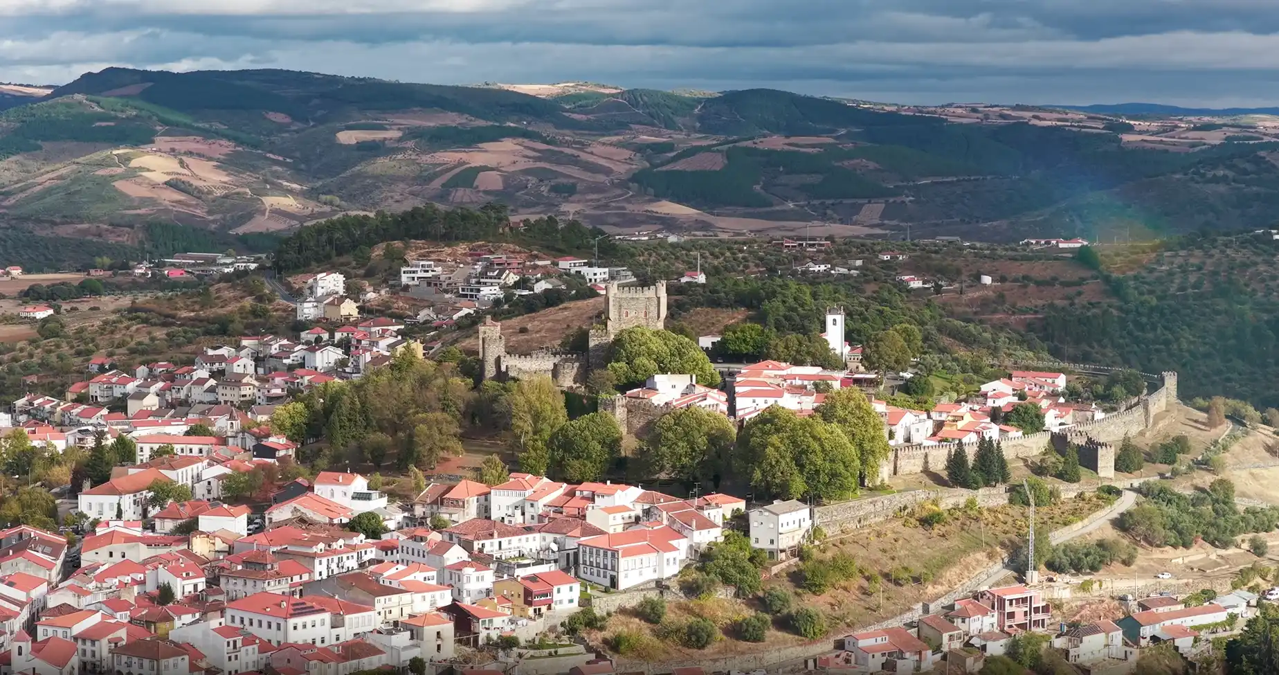 Vista a&eacute;rea de uma vila com um castelo e colinas ao fundo.