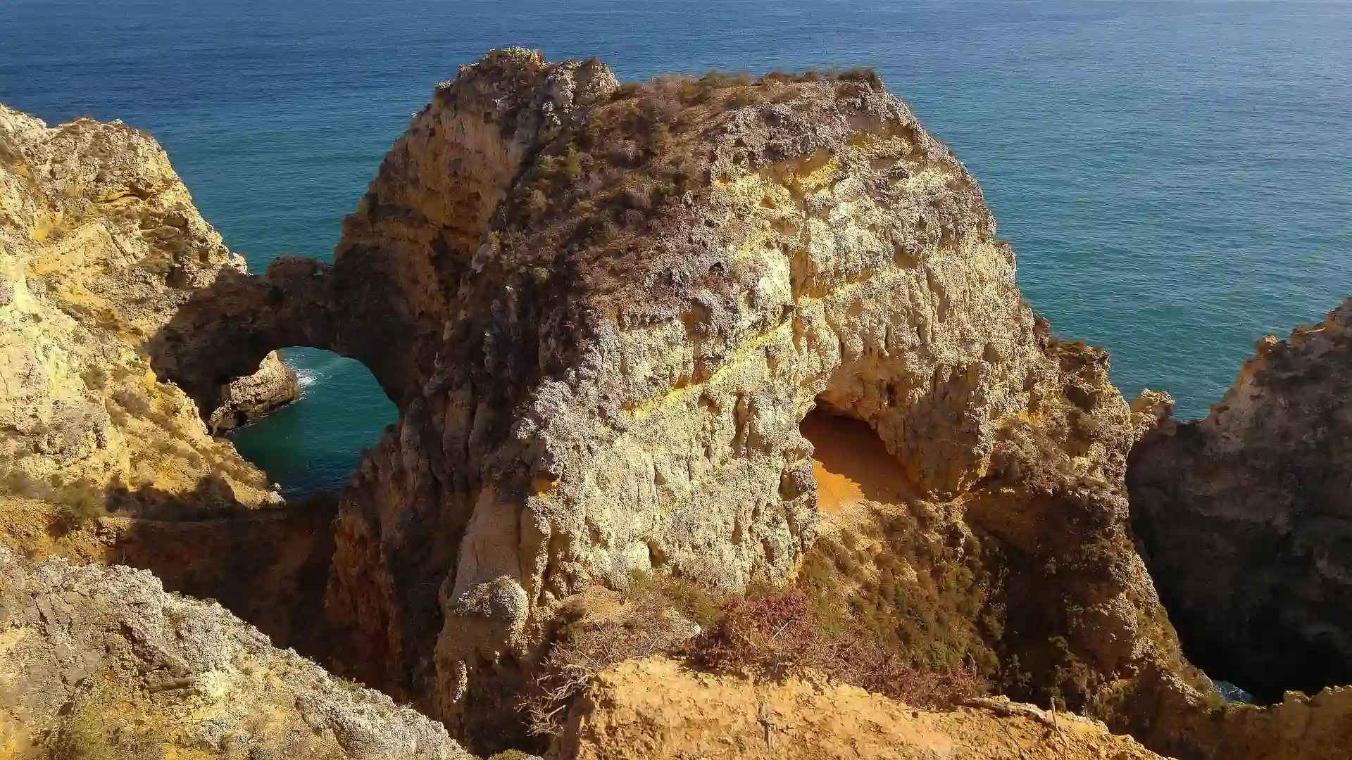 Forma&ccedil;&atilde;o rochosa &agrave; beira-mar com arcos naturais e &aacute;guas azuis ao fundo.