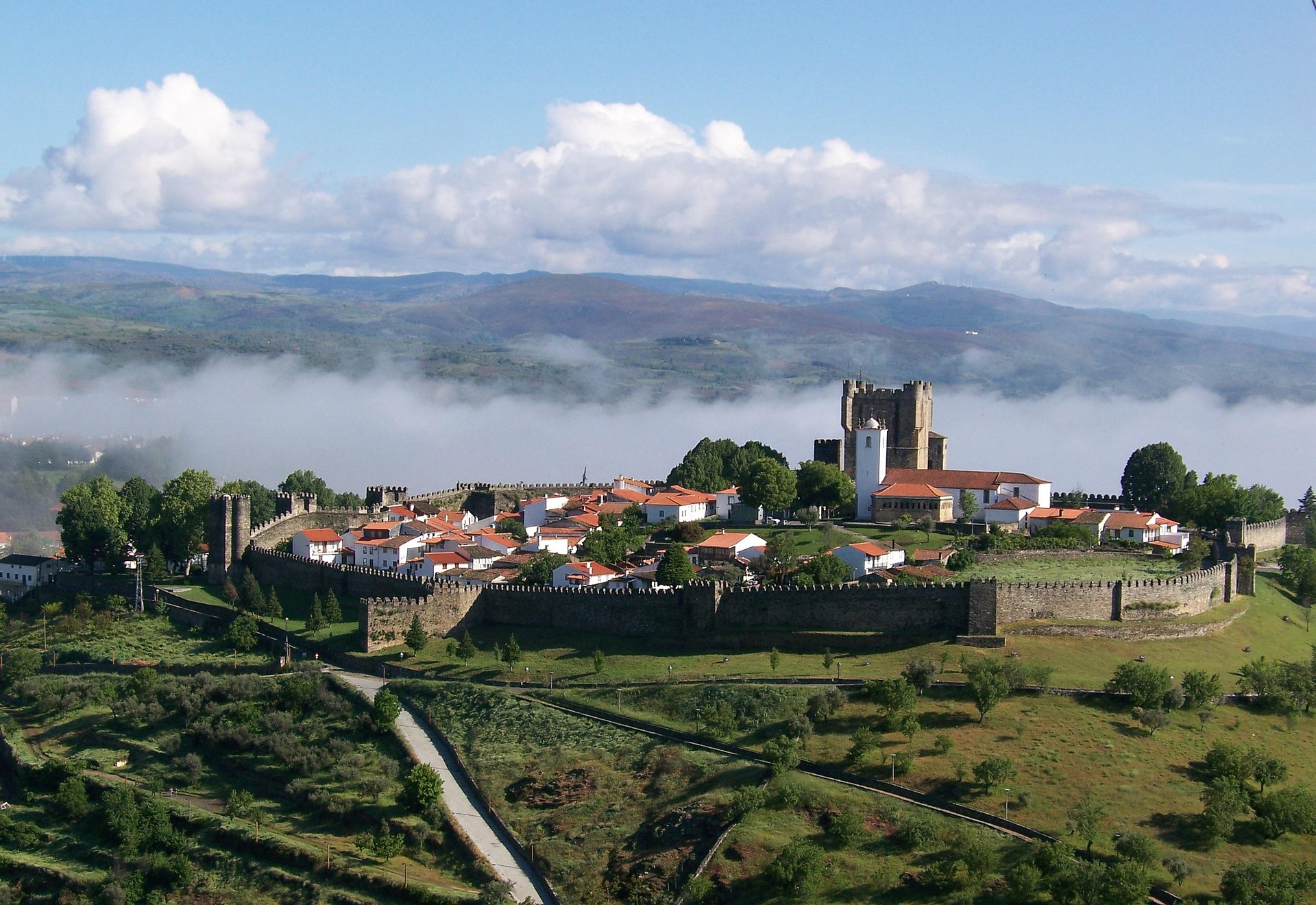Castelo hist&oacute;rico cercado por muralhas com montanhas ao fundo.