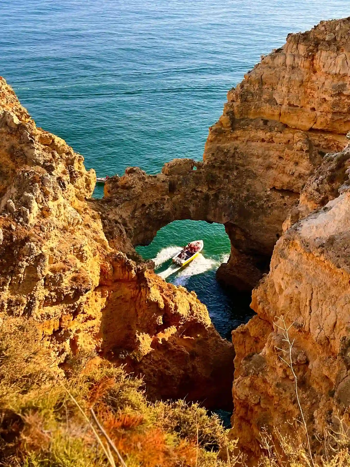 Barco passa por arco entre fal&eacute;sias rochosas com mar azul ao fundo.