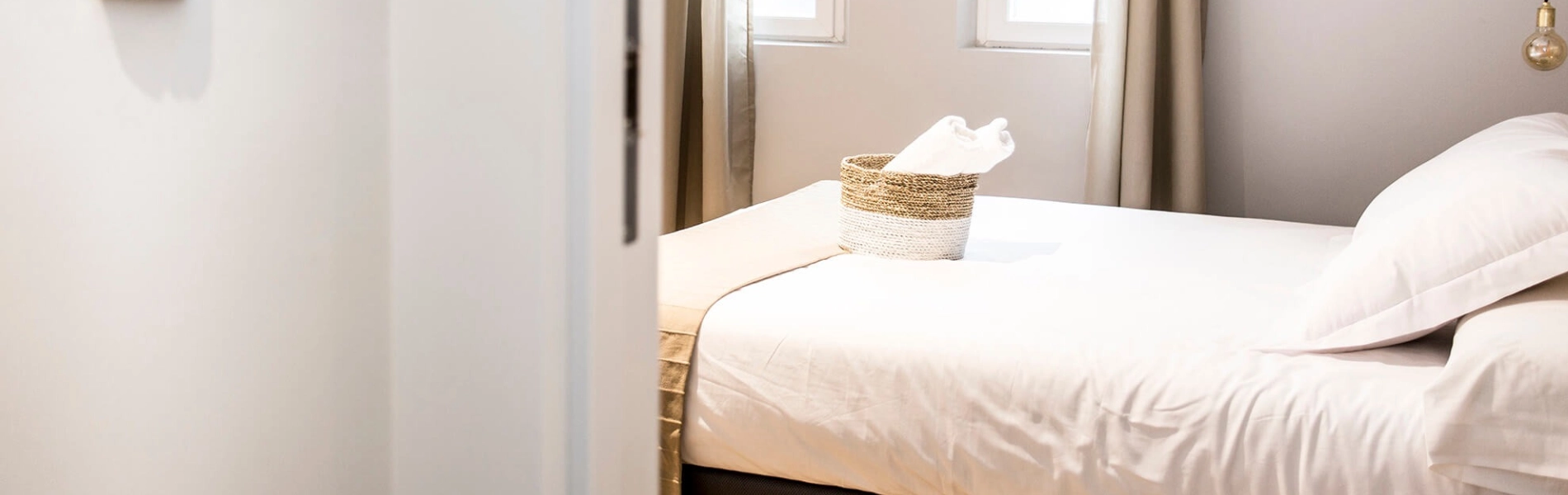 Habitaci&oacute;n luminosa con cama hecha y cesta de toallas.