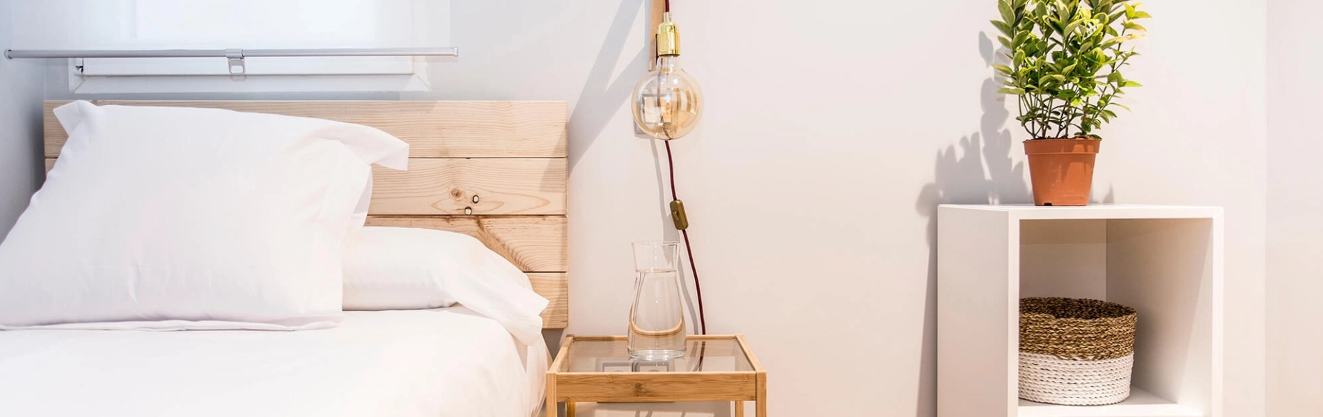 Habitaci&oacute;n luminosa con cama, l&aacute;mpara, planta y estante al lado.