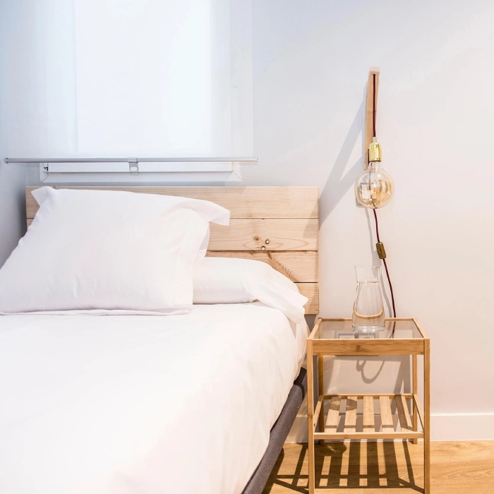 Cama con cabecera de madera, mesa con jarra y bombilla colgante blanca.