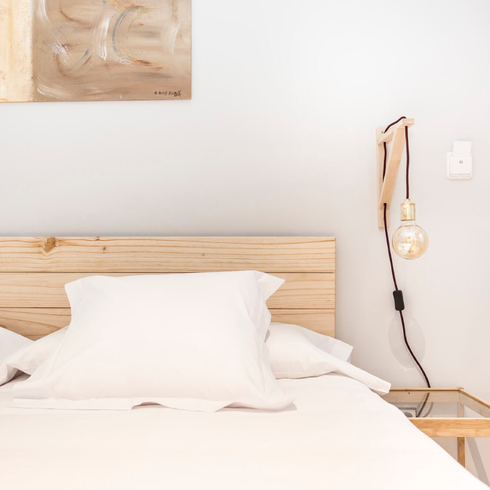 Cama minimalista con cabecero de madera y l&aacute;mpara colgante en pared blanca.