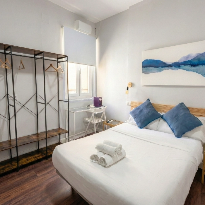 Habitaci&oacute;n con cama, estanter&iacute;a, cuadro azul y ventana.