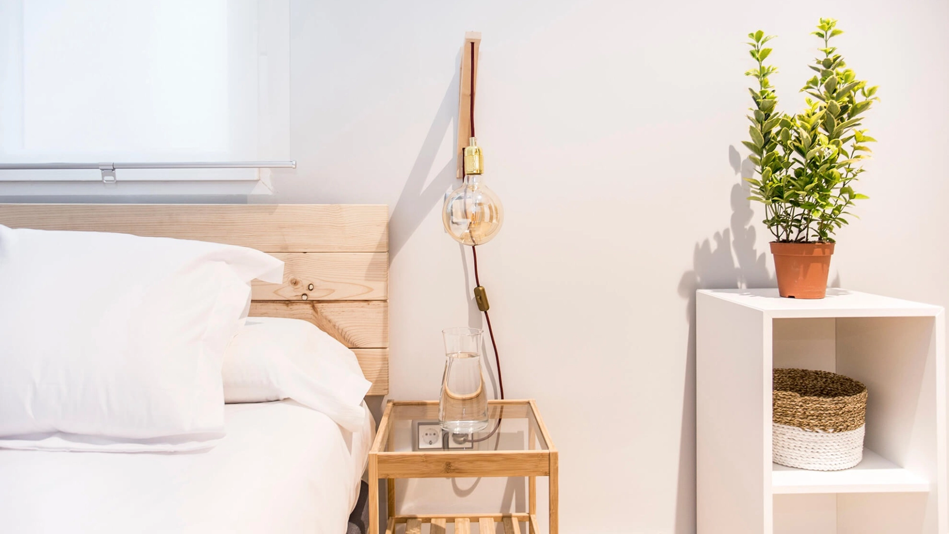 Cama con l&aacute;mpara colgante, mesa con jarra y repisa con planta en maceta.