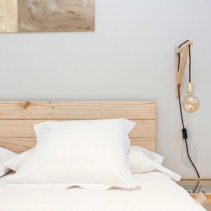 Cama con cabecera de madera, l&aacute;mpara colgante y cuadro en la pared.