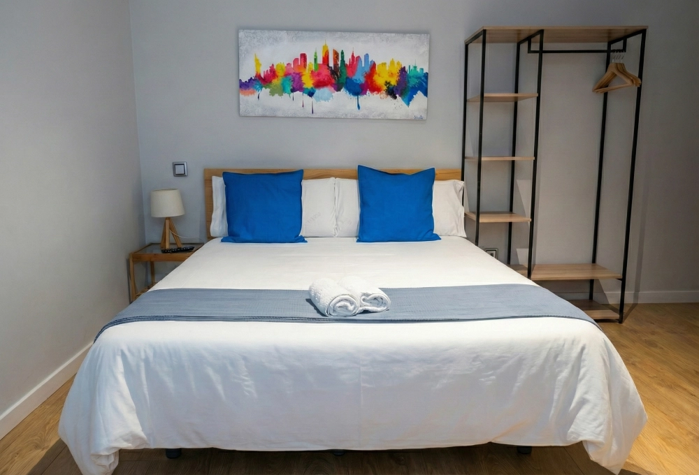 Cama con cojines azules, estante y cuadro colorido sobre pared blanca.