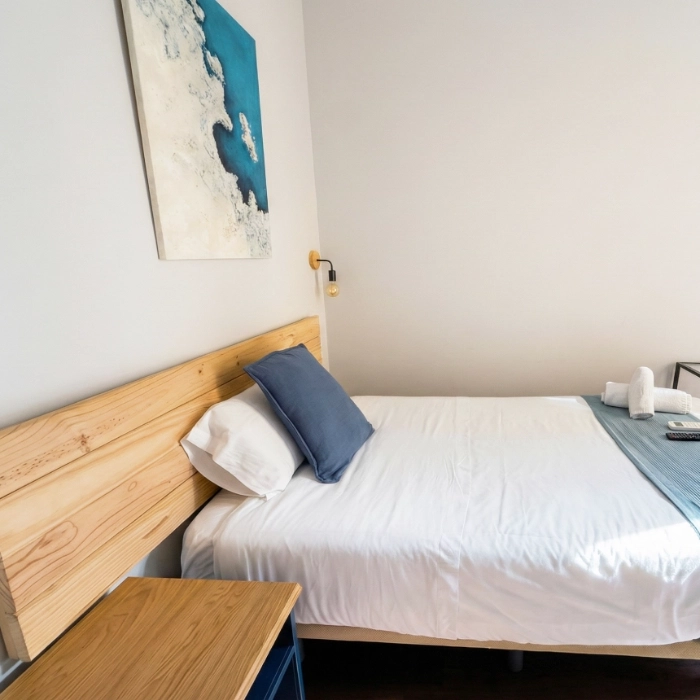 Cama simple en habitaci&oacute;n con cuadro azul y mesa peque&ntilde;a de madera.