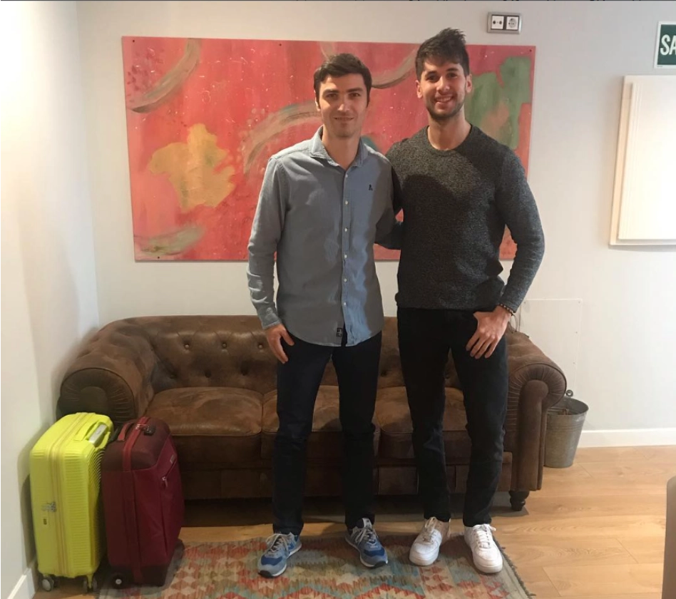 Dos hombres posan frente a un sof&aacute; con maletas al lado y un cuadro detr&aacute;s.