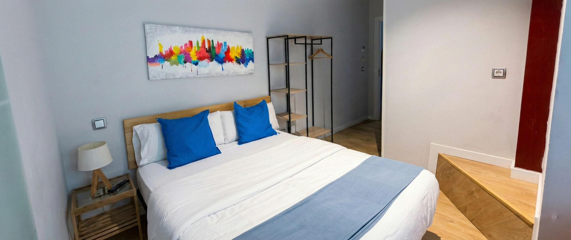 Habitaci&oacute;n moderna con cama doble, cojines azules y cuadro colorido en la pared.