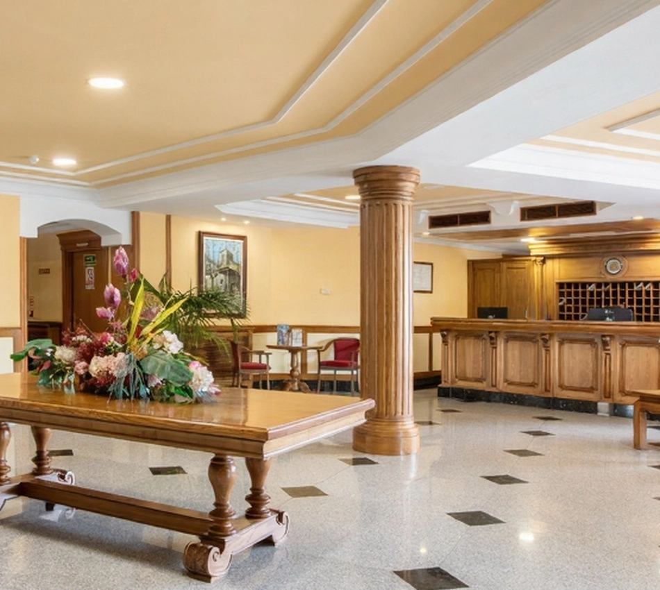 Vest&iacute;bulo de hotel elegante con mesa de centro, flores y recepci&oacute;n al fondo.