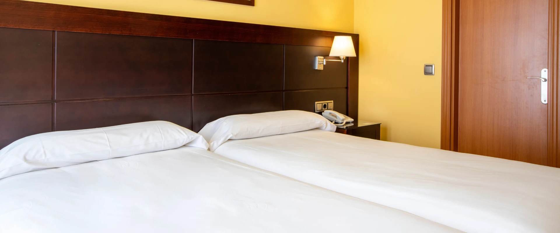 Cama doble con cabecera de madera y l&aacute;mpara de pared en habitaci&oacute;n amarilla.