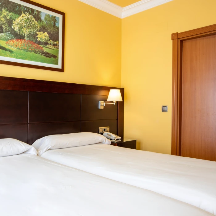 Cama doble en habitaci&oacute;n amarilla con l&aacute;mpara y cuadro de paisaje en la pared.