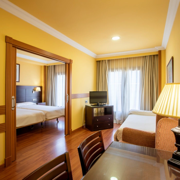 Habitaci&oacute;n de hotel con tres camas, mesa y televisi&oacute;n sobre mueble.