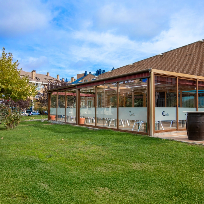 Terraza acristalada en un jard&iacute;n, con sillas y mesas. Cielo despejado.