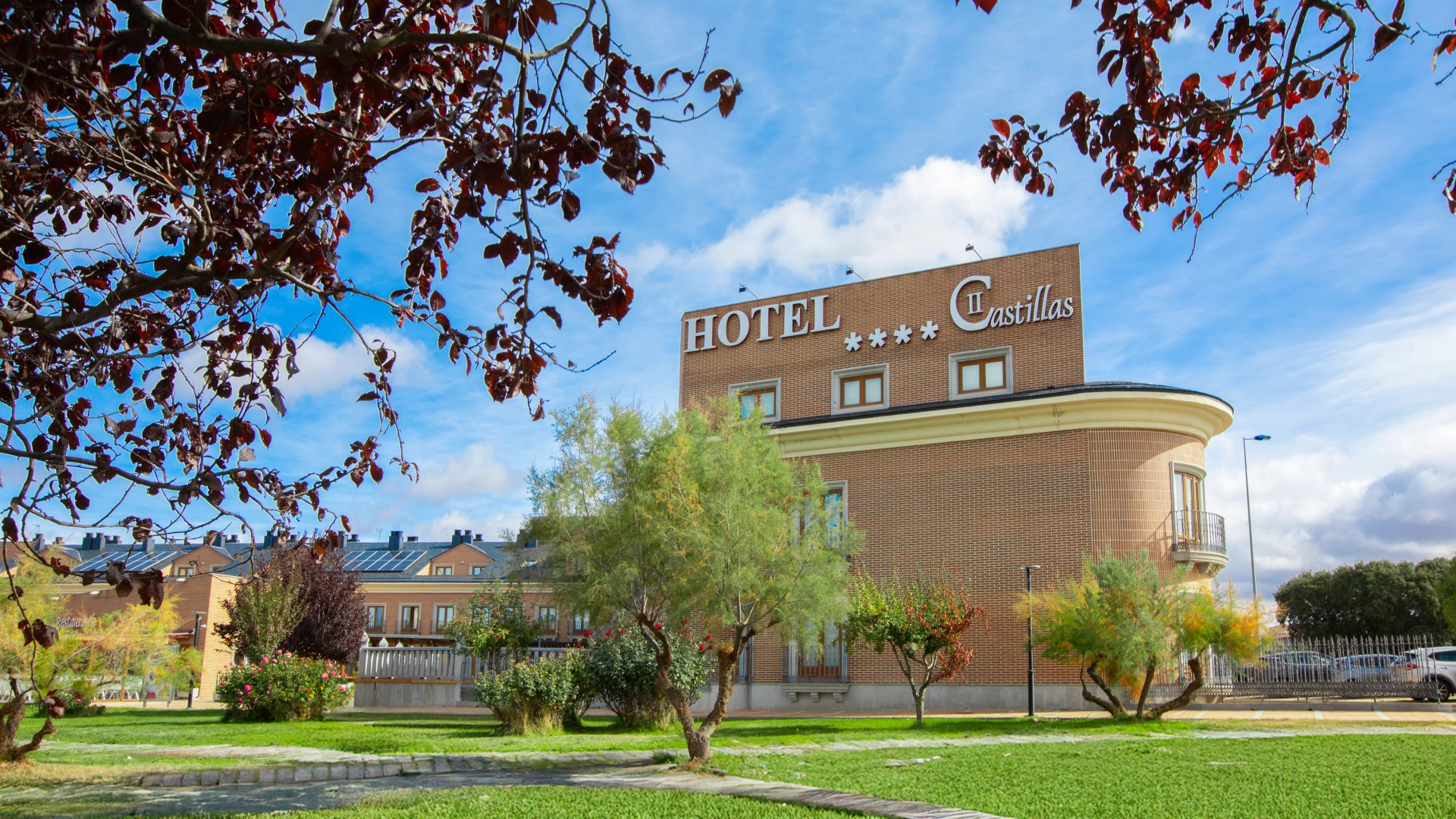Edificio del Hotel II Castillas rodeado de &aacute;rboles y c&eacute;sped bajo un cielo azul.