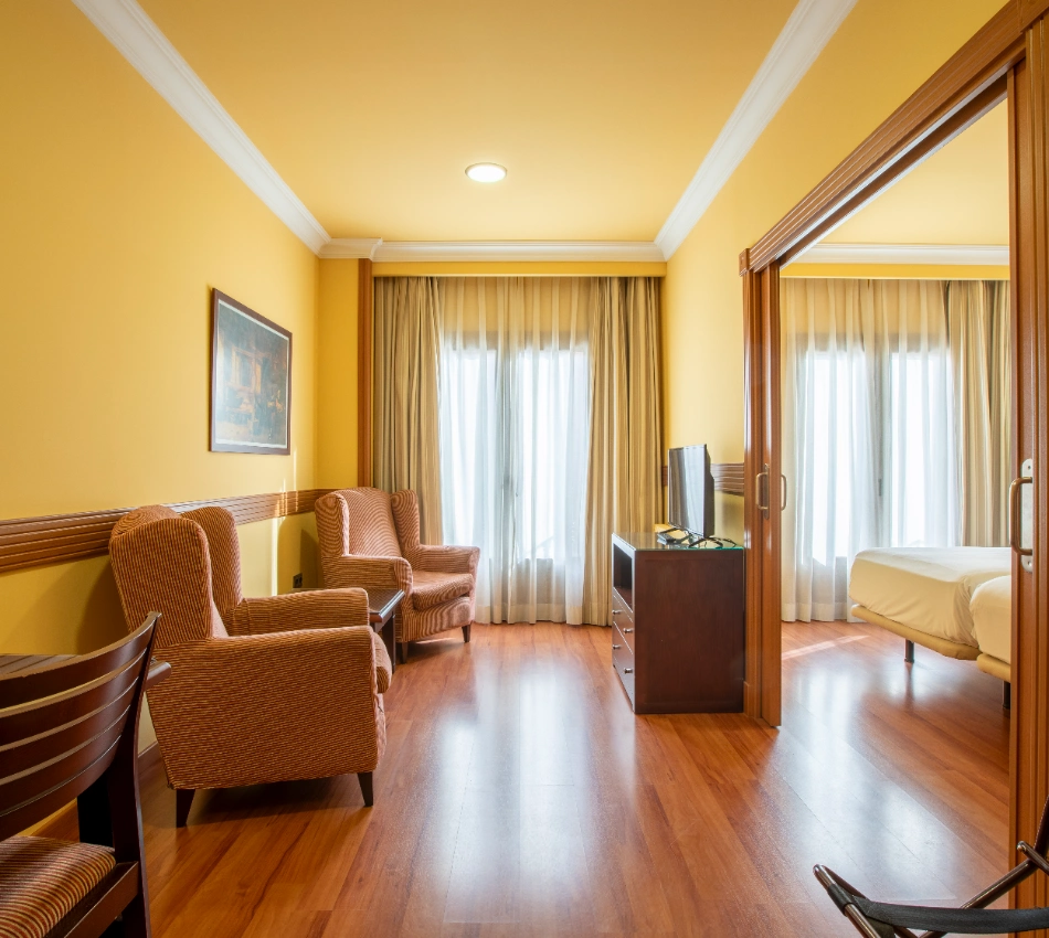 Sala de hotel con sillones, TV y ventana, conectada a un dormitorio.