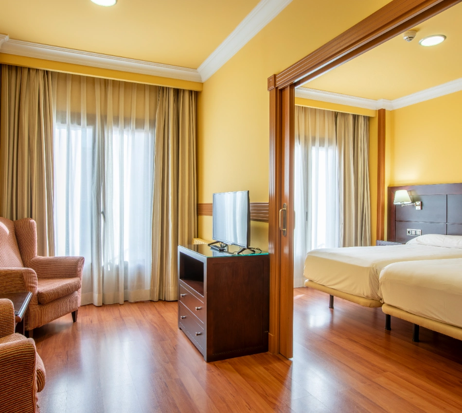 Habitaci&oacute;n de hotel con camas, sillones y televisor, decorada en tonos c&aacute;lidos.