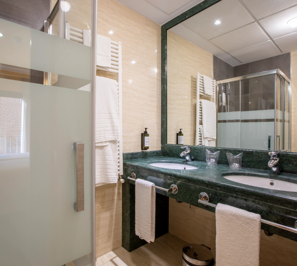 Ba&ntilde;o moderno con lavabo doble, toallas y ducha con puerta de vidrio.