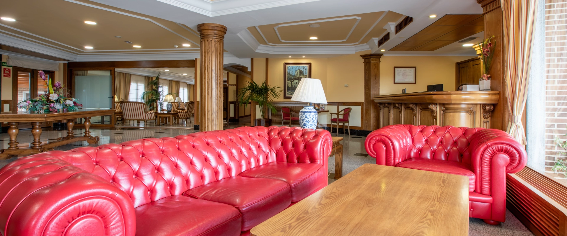 Sala de estar con sof&aacute;s rojos, mesa de madera y recepci&oacute;n al fondo.