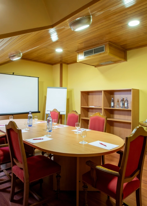 Sala de reuniones con mesa ovalada y sillas rojas, pantalla y pizarra.