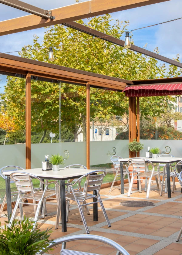 Terraza con mesas de metal, plantas y techo de madera en un d&iacute;a soleado.
