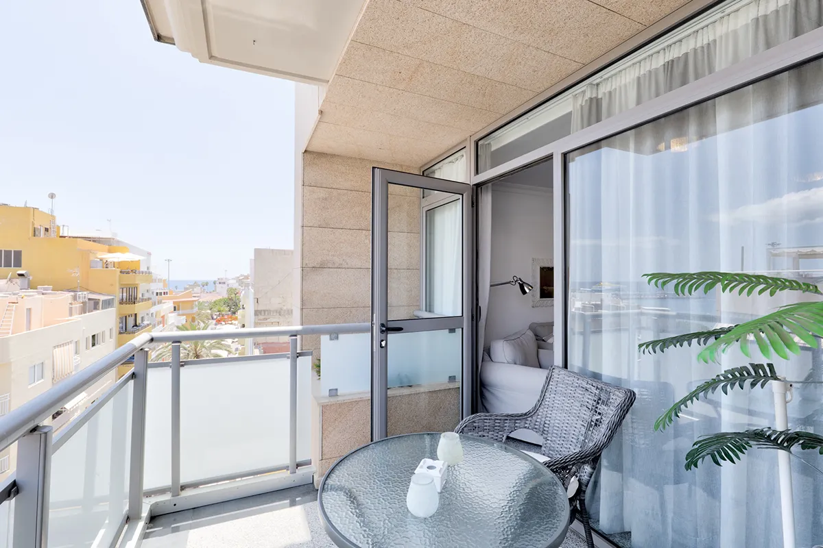 Balcón moderno con mesa y silla de mimbre, vista urbana y planta decorativa.