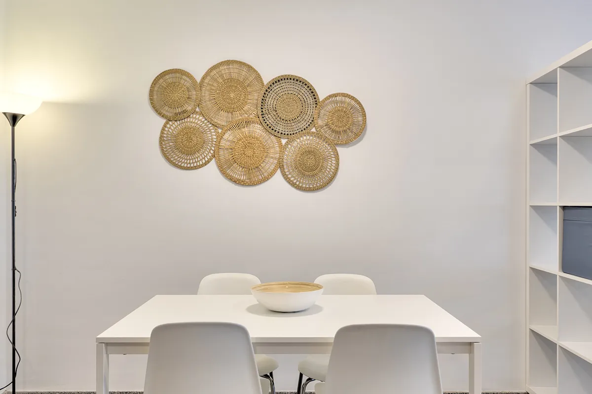 Mesa blanca con sillas, pared decorada con cestas y lámpara de pie.