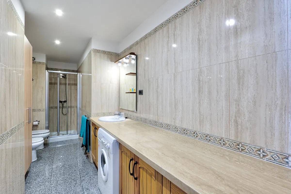 Baño moderno con ducha, lavamanos, lavadora y azulejos beige decorativos.