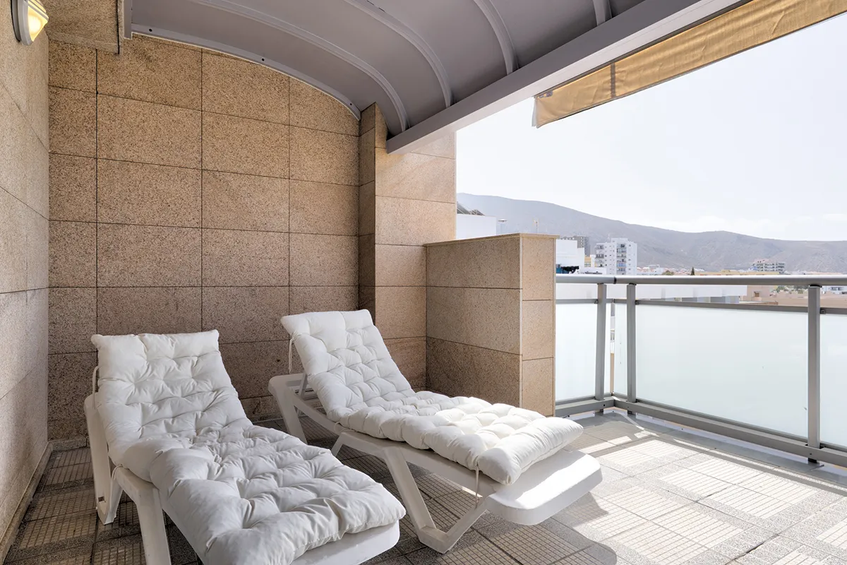 Dos tumbonas blancas en un balcón con vista a la ciudad y montañas.